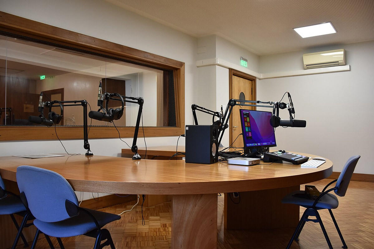 A purpose-built podcast studio with microphones on boom arms and a control-room window